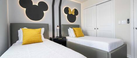 Ein Hotelzimmer mit zwei Betten, jeweils mit Kopfstütze und Kissen. An der Wand befinden sich Mickey-Mouse-Dekorationen, und die Tür ist mit einem Zahlenschloss versehen.