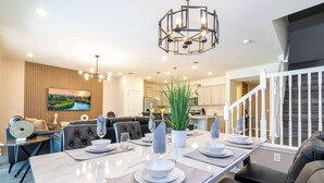 A modern dining area with a long table set for four, a chandelier, and a view of the kitchen and living area in the background.