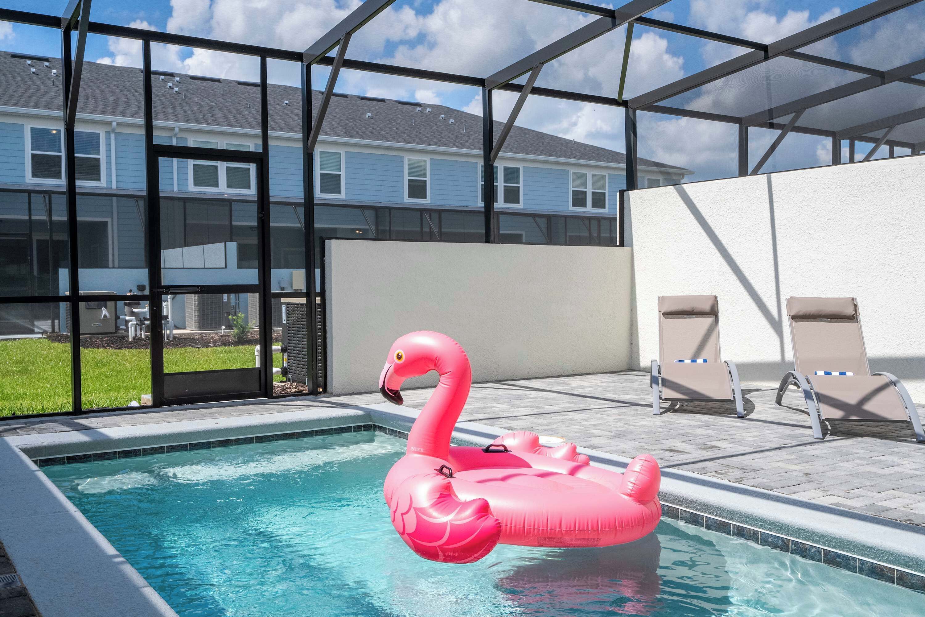 A swimming pool with a pink flamingo float, a glass-enclosed patio with lounge chairs, and a view of a residential building.