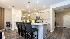 A modern kitchen with a central island, bar stools, pendant lighting, and a dining area with a high chair.
