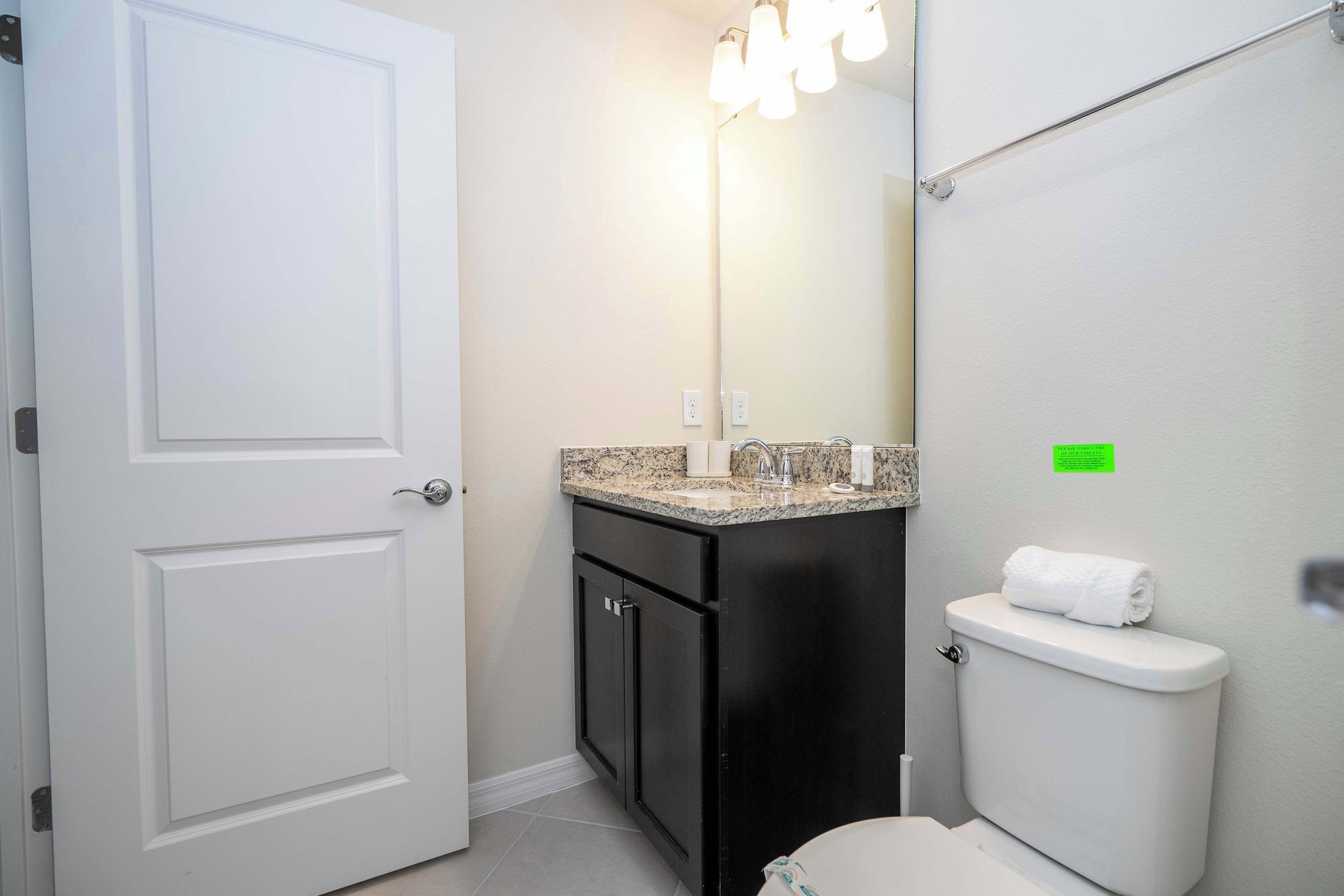 A bathroom with a white door, a sink with a granite countertop, a toilet, and a mirror with lights above it.