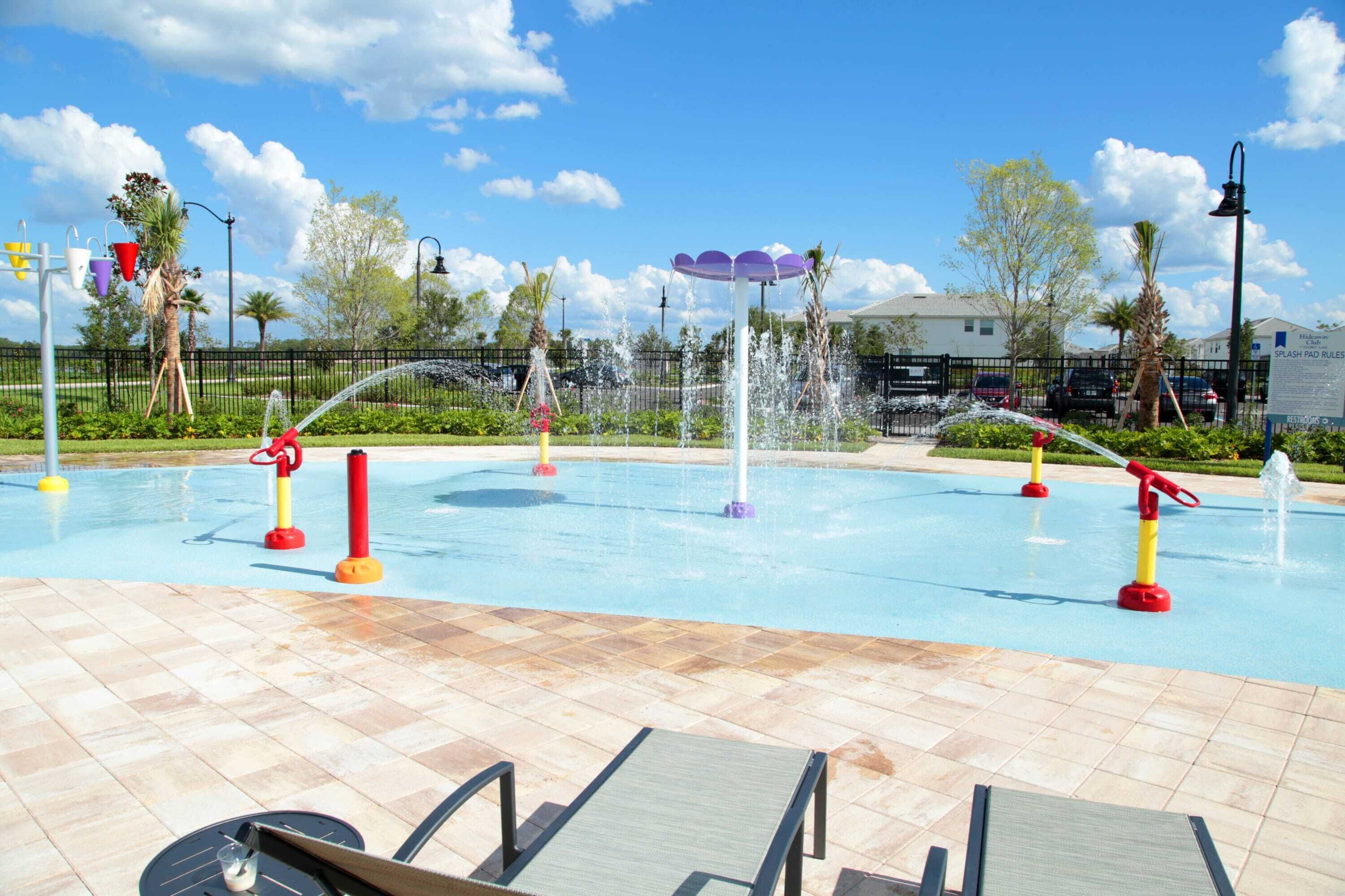 Una piscina con múltiples chorros de agua, rodeada por un deck empedrado y palmeras.