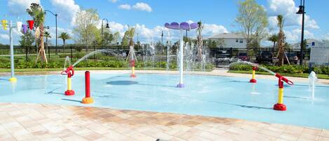 Una piscina con múltiples chorros de agua, rodeada por un deck empedrado y palmeras.