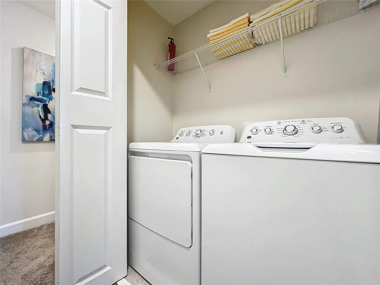A laundry room with two stacked front-loading washers and dryers, a fire extinguisher on the wall, and a shelf above holding folded towels.