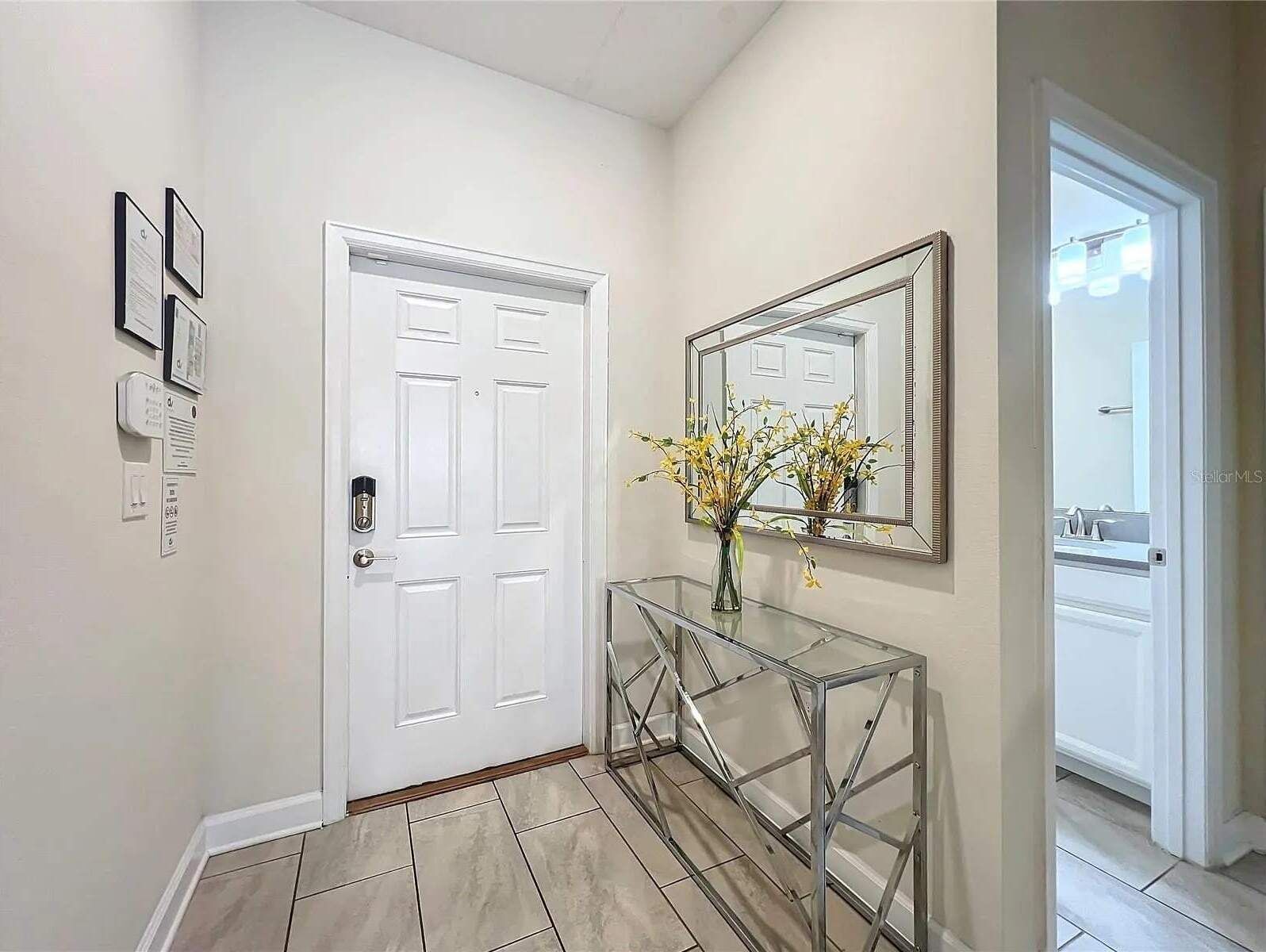 A hallway with a white door, a glass console table with a vase of flowers, and a mirror on the wall.