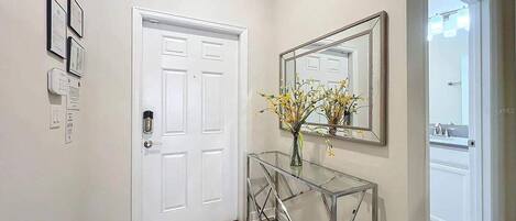 A hallway with a white door, a glass console table with a vase of flowers, and a mirror on the wall.
