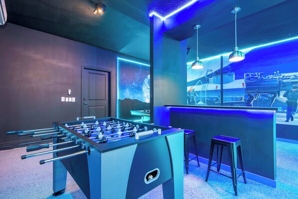 A room with a foosball table, a bar area with neon lights, and a seating area with a stool. - Stunning 7Bd Jacuzzi Pool & Close To Disney @ Windsor Cay 16217 (Clermont)