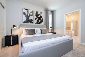 Une chambre moderne avec un grand lit, des tables de chevet avec des lampes, des œuvres d’art abstraites accrochées au mur et une salle de bain visible à travers une porte ouverte.