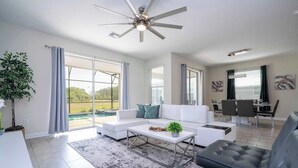 A modern living room with a white sofa, a black leather couch, a coffee table, and a ceiling fan. There is a dining area with a table and chairs, and a large window with curtains. - Stunning Game Room & Pool Jacuzzi Storey Lake 2799 (Kissimmee)
