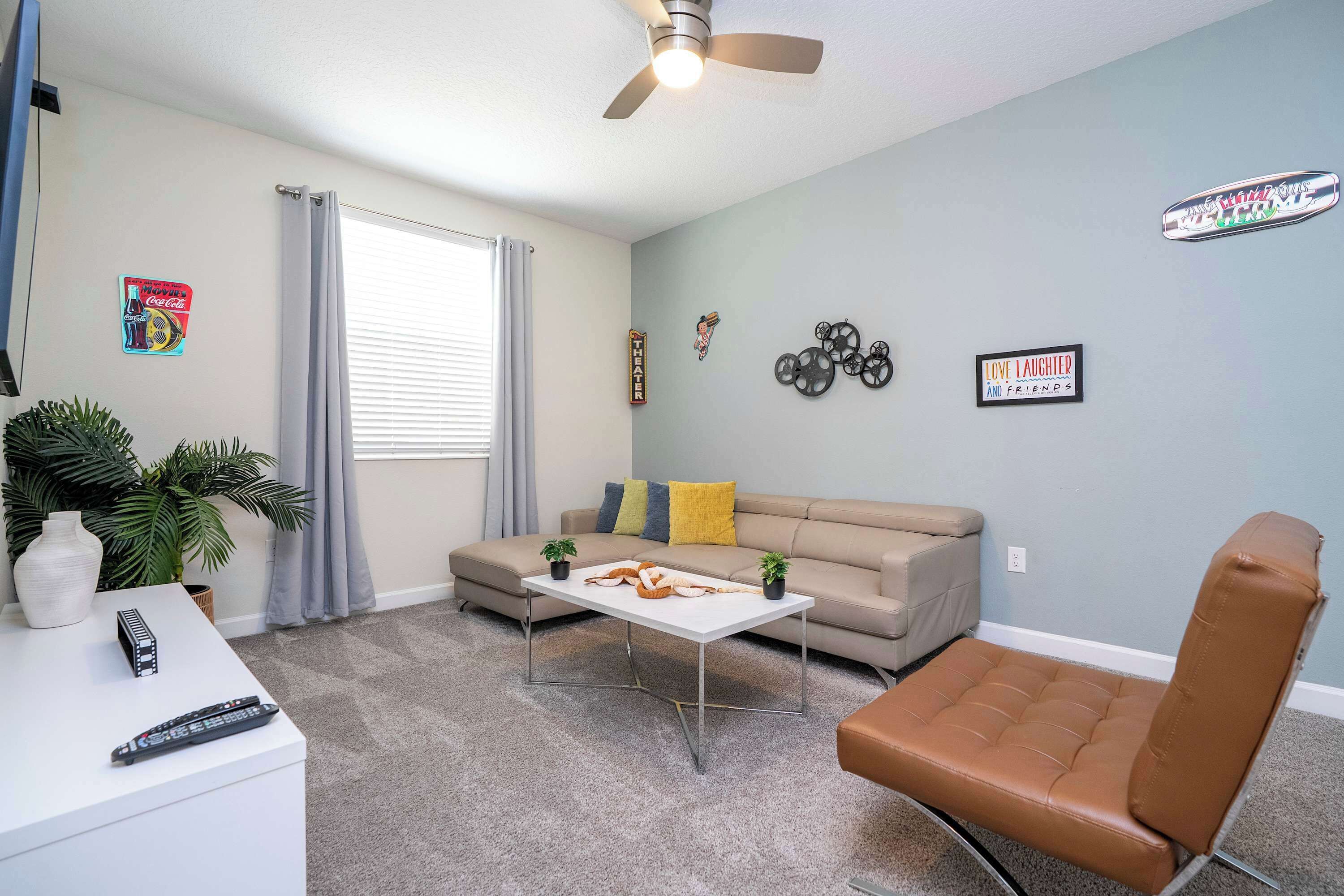 Un salon comprenant un canapé, une table basse, un fauteuil en cuir marron, un ventilateur de plafond et une fenêtre avec des rideaux.