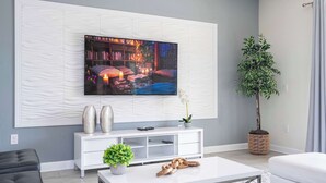 A modern living room with a flat-screen TV mounted on a textured wall, a white console table with decorative items, a coffee table with shoes, and a potted plant. - Stunning Game Room & Pool Jacuzzi Storey Lake 2799 (Kissimmee)