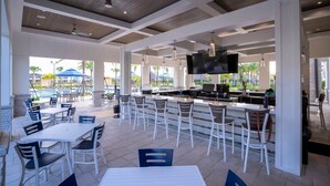An outdoor bar area with white tables and chairs, a flat-screen TV mounted above the bar, and a pool visible in the background. - Wonderfull 4Bd W/ Pool @ Champions Gate Resort 8945 (Davenport)