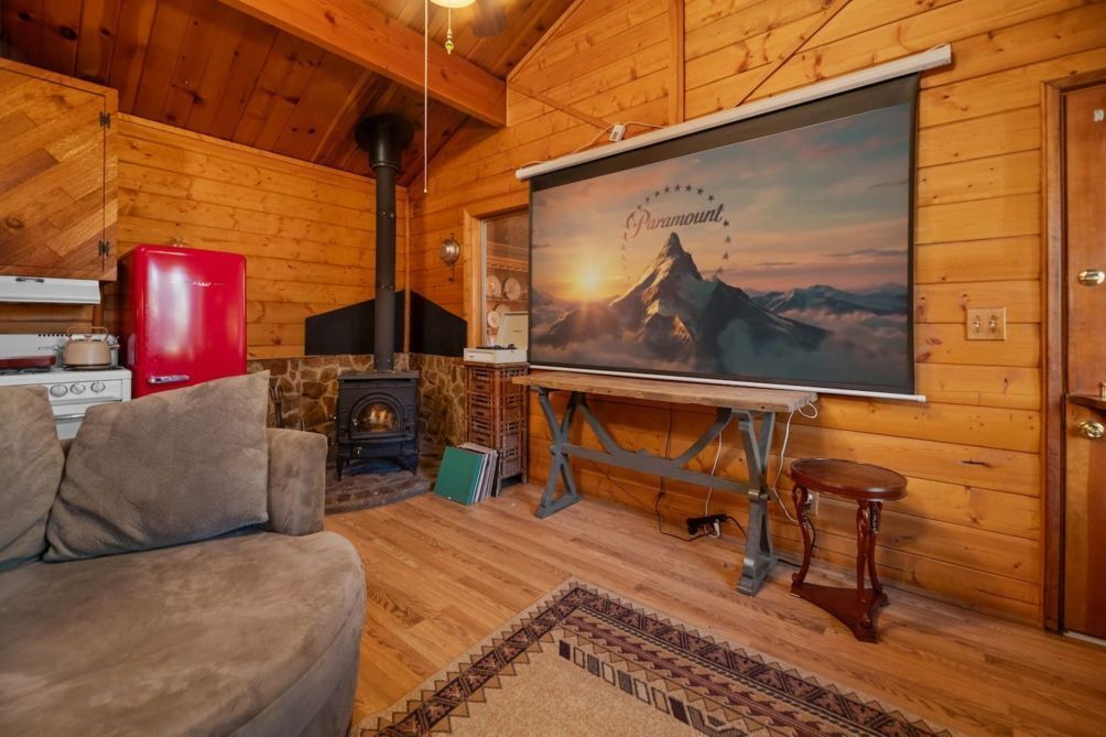 Ein gemütliches Blockhaus-Interieur mit Holzausstattung, Kamin, einem roten Kühlschrank und einer an der Wand montierten Projektionsleinwand, die eine Berglandschaft zeigt.