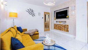 A living room with a yellow sofa, a TV mounted on the wall, a lamp, and a fireplace. - Driveway | Modern Home In Altrincham | Sleeps 7 (Altrincham)