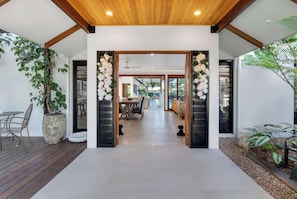 A modern interior with a wooden ceiling, a central doorway flanked by white flowers, a dining area with a table and chairs, and a view into the adjacent room.