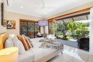 A bedroom with a bed, pillows, a TV, a ceiling fan, and a view of the outdoors.
