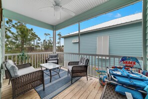 Relax in a serene outdoor space with a wooden deck, wicker chairs, and a ceiling fan, surrounded by trees and a charming building.