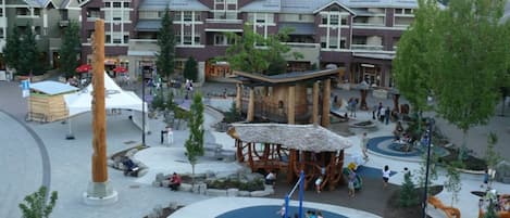 A playground with a blue circular area, surrounded by a plaza with benches and trees.