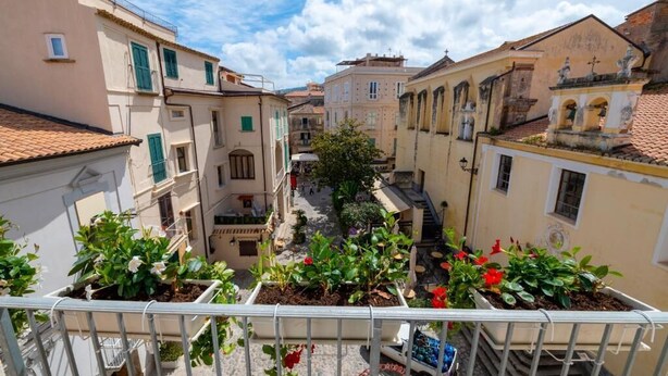 Une rue étroite, bordée de bâtiments des deux côtés, avec des balcons ornés de plantes en pot et un ciel dégagé.