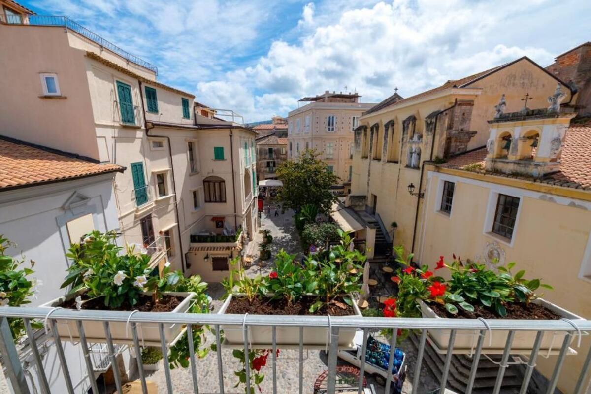 Rua estreita com edifícios em ambos os lados, varandas com plantas em vasos e um céu claro.