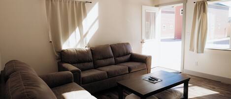 A living room with a brown leather sofa, a wooden coffee table, and a view of a building through an open door.