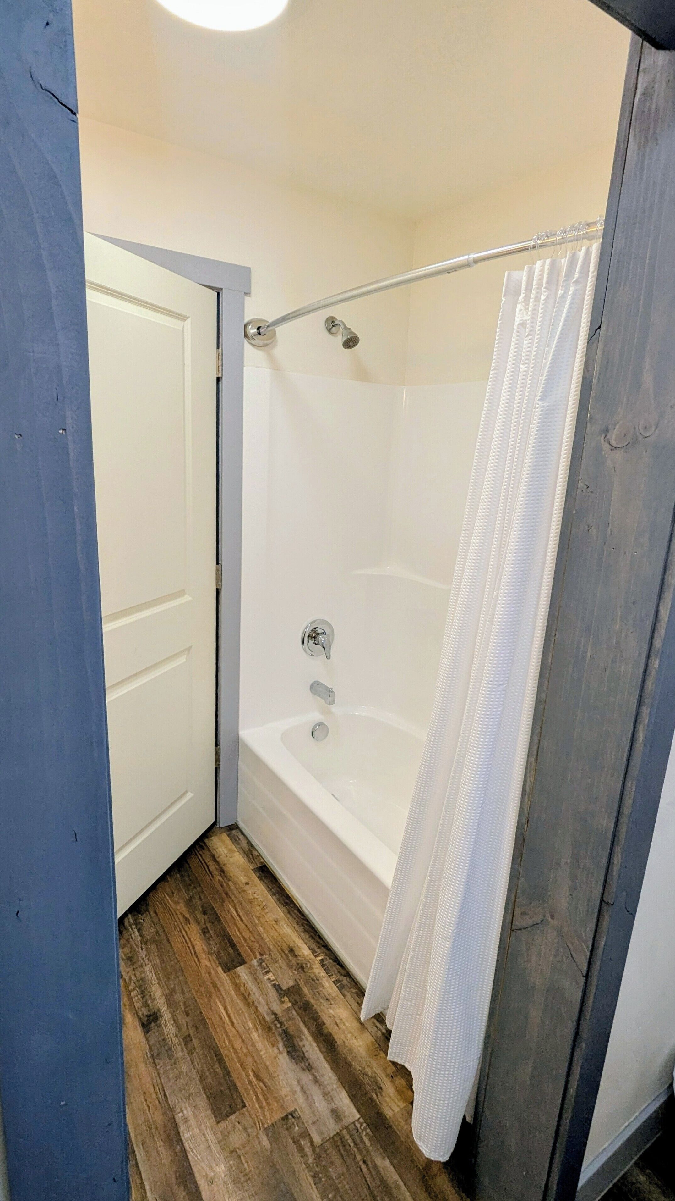 A small, enclosed shower area with a bathtub, wooden flooring, and a white shower curtain.