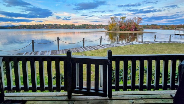 Eine Holzterrasse mit Blick auf einen See, einen Steg und herbstlich gefärbte Bäume.