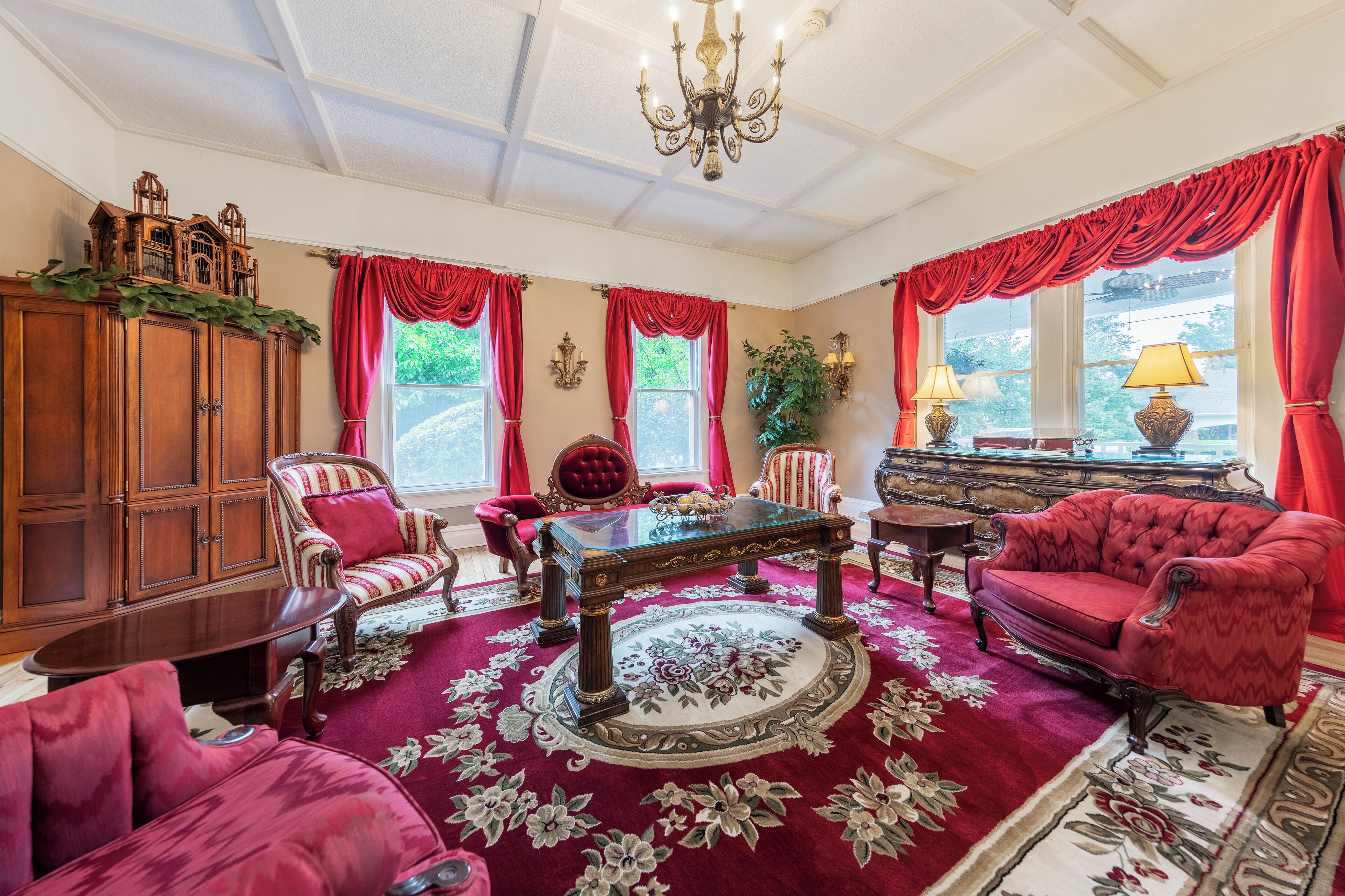 A room with a wooden cabinet, a glass-top table, red upholstered chairs, and a red carpet with floral patterns.