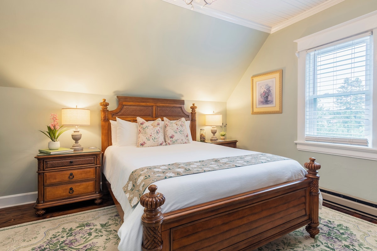 A bedroom with a wooden bed, two bedside tables with lamps, a floral rug, and a window with blinds.