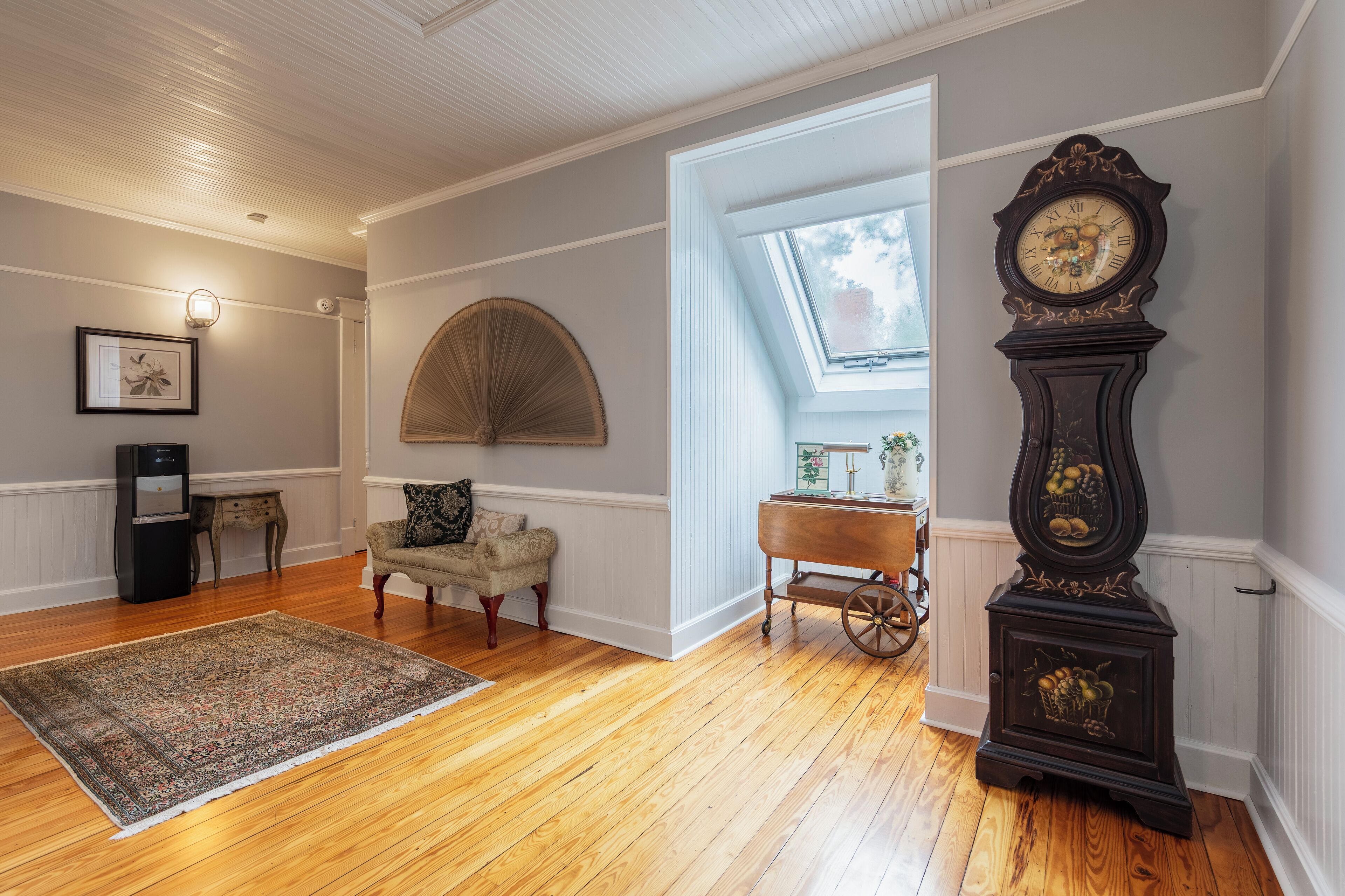 Une pièce avec un plancher en bois, une grande moquette, une horloge ancienne, un ventilateur et une petite table avec une lampe.