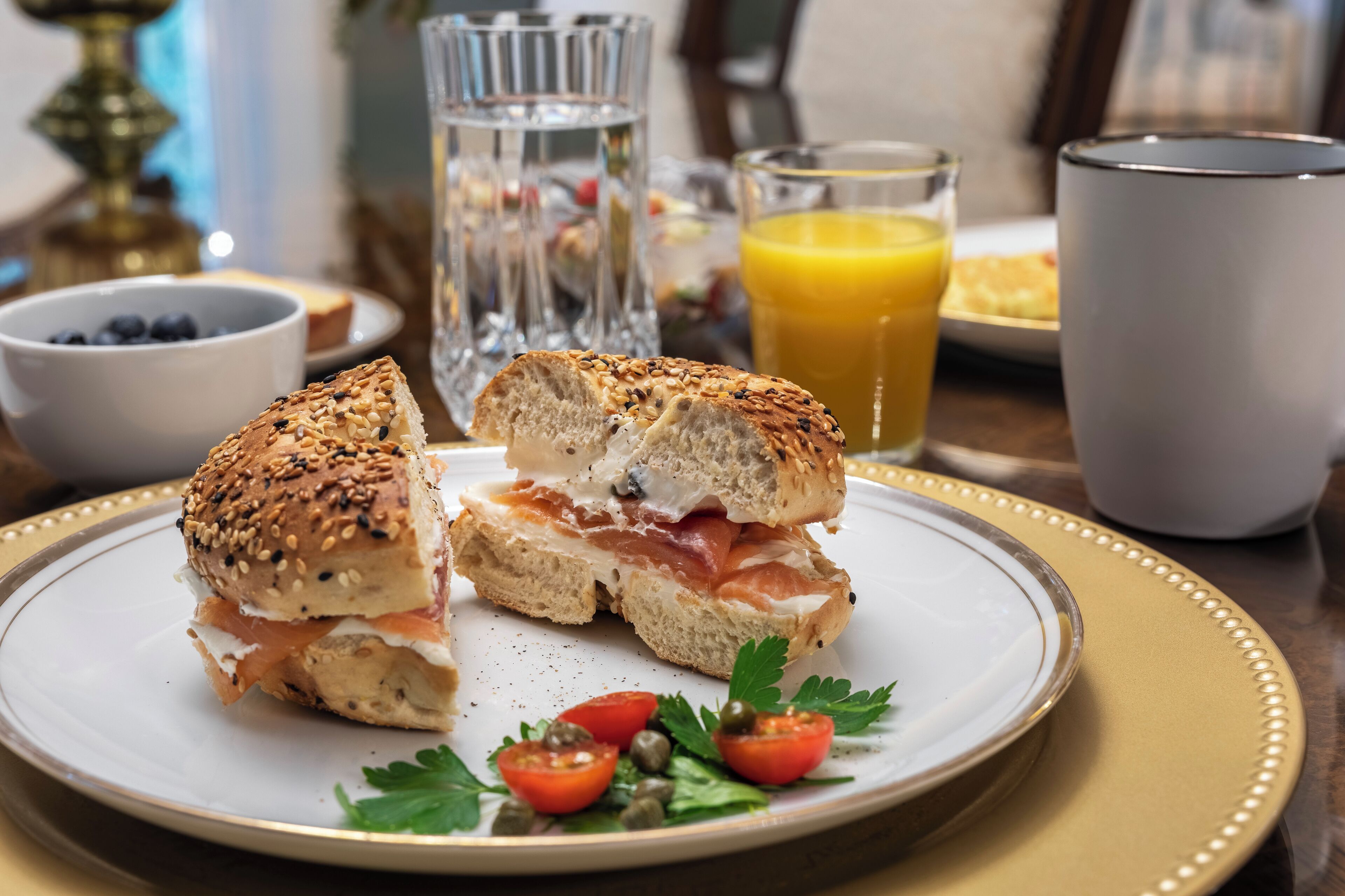 A breakfast sandwich on a plate with a side of tomatoes and capers, a glass of orange juice, and a cup of coffee.