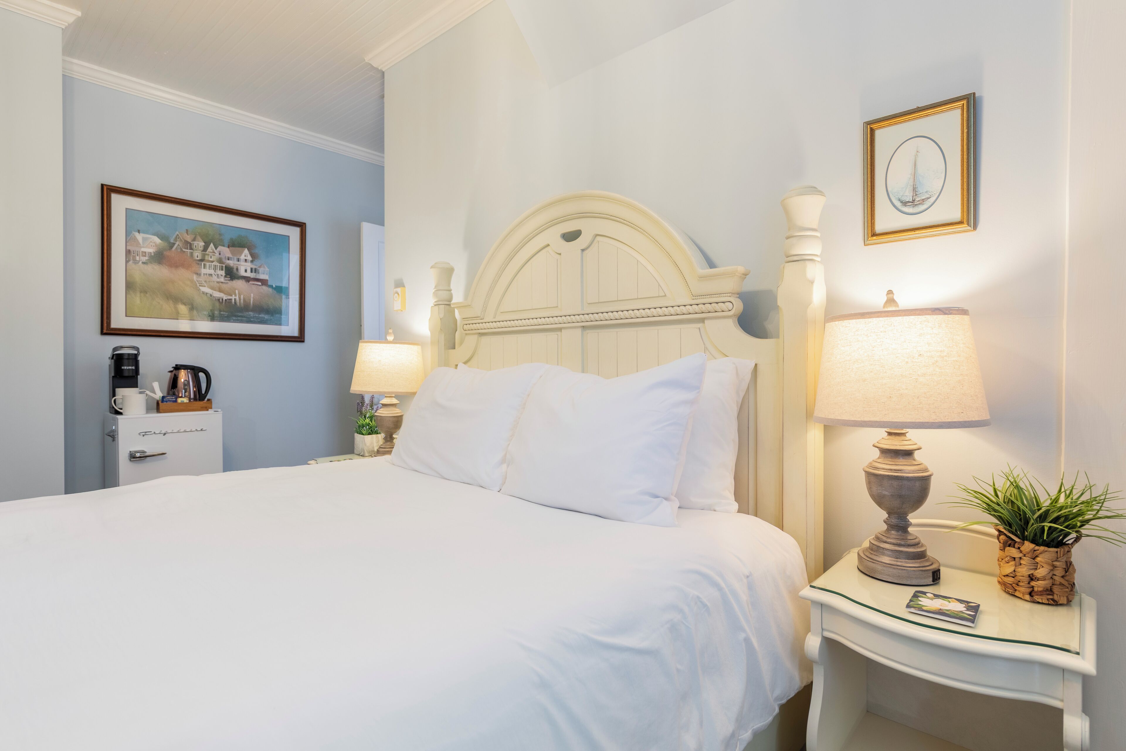 A neatly made bed with white linens, a bedside table with a lamp and a small potted plant, and a framed picture on the wall.