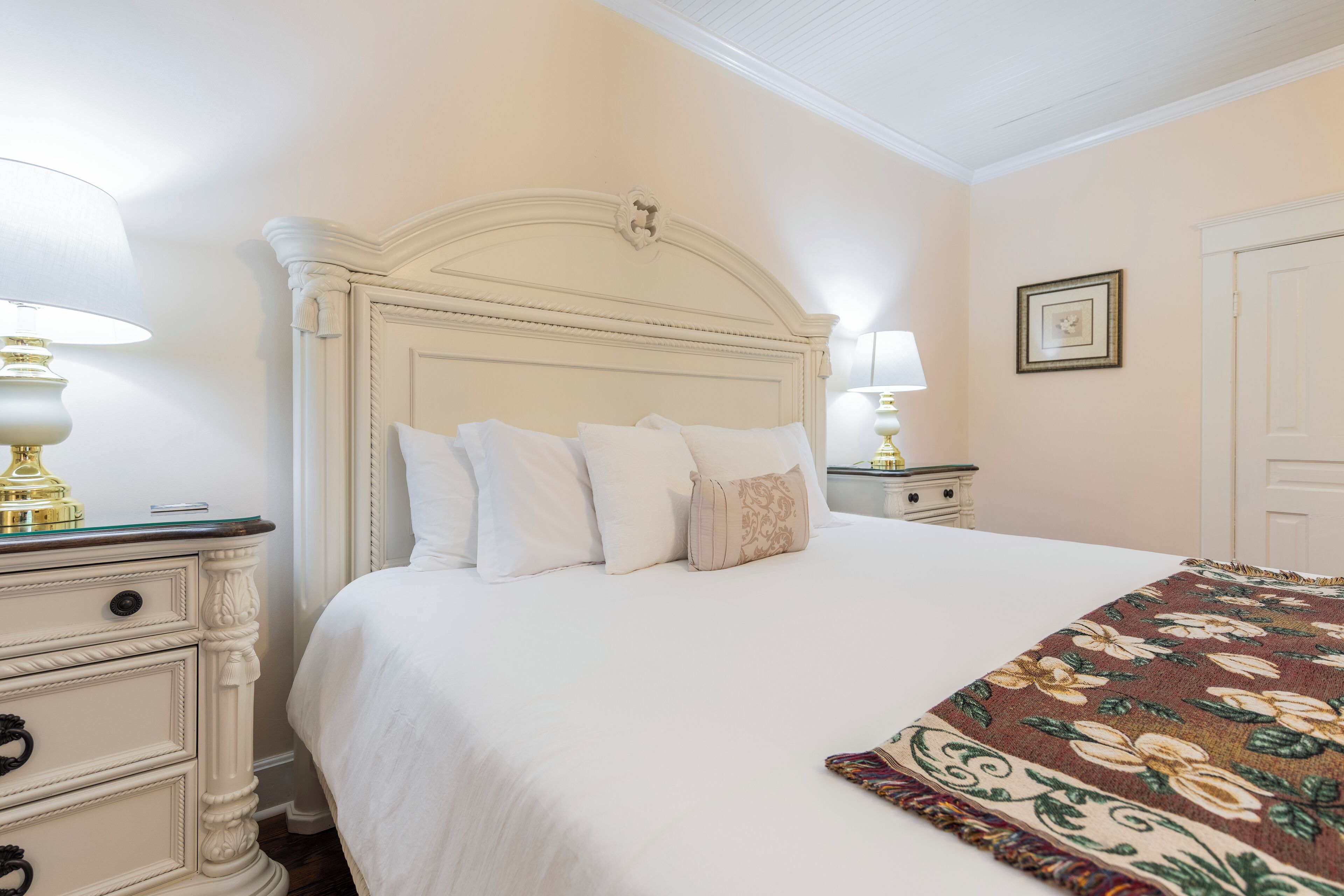 A neatly made bed with white linens and a floral throw blanket, a white nightstand with a lamp, and a framed picture on the wall.