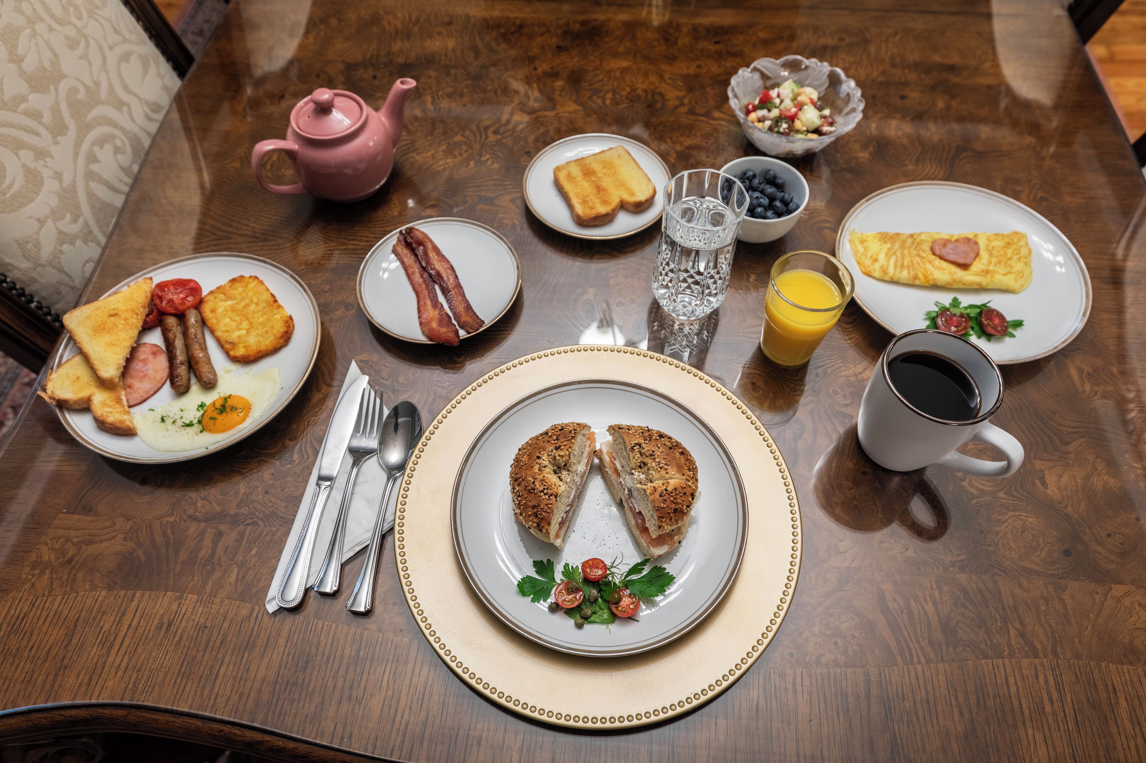 Una mesa de desayuno con diversos platos, como tostadas, huevos, tocino y un bagel, junto con un hervidor de agua, un vaso de agua, un vaso de jugo de naranja y una taza de café.