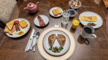 Una mesa de desayuno con diversos platos, como tostadas, huevos, tocino y un bagel, junto con un hervidor de agua, un vaso de agua, un vaso de jugo de naranja y una taza de café.
