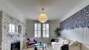 Una sala de estar con pared de piedra, chimenea, un sillón púrpura, sofá blanco, una mesa de centro de vidrio y una lámpara colgante moderna.