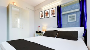 A hotel room with a large bed, a nightstand, a desk, and a window with blue curtains. - Qubenest Splendour (Bangalore)