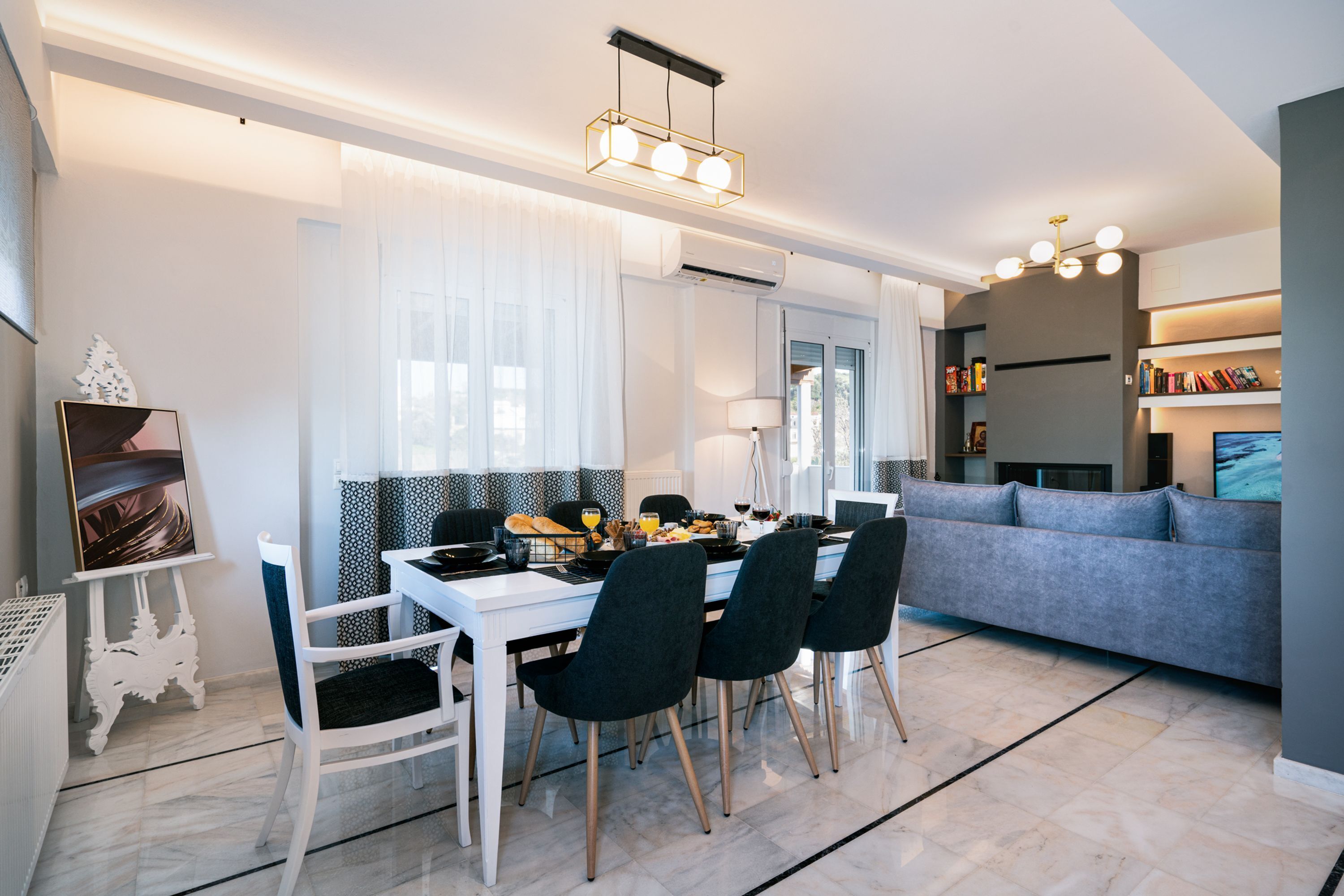 A modern living room with a grey sofa, a dining area with a white table and black chairs, and a kitchen area with a white countertop and a hanging light fixture.