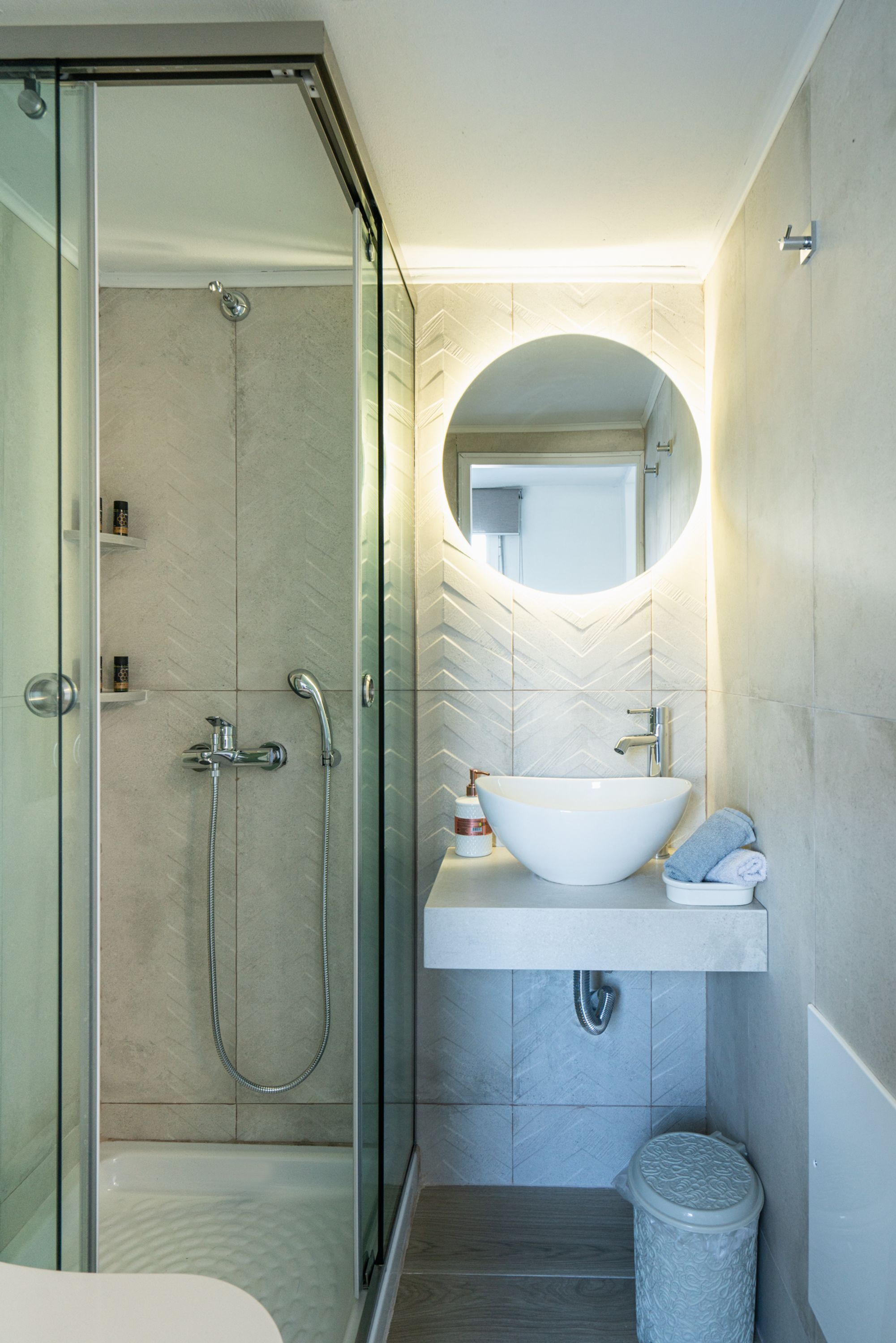 Une salle de bain moderne avec une douche à l’italienne en verre, un lavabo blanc avec un receveur et un miroir rond au-dessus du lavabo.