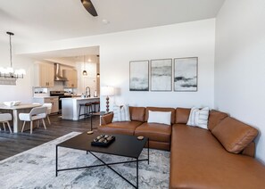 Una sala de estar moderna con un sofá de cuero marrón, una mesa de centro y un comedor con una mesa y sillas blancas. Se observa una cocina al fondo con armarios blancos y una isla central.
