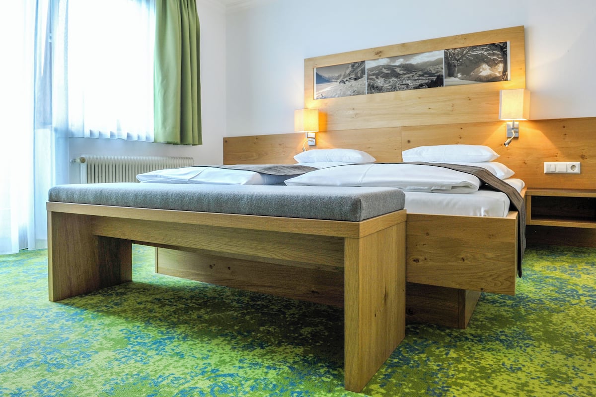 A hotel room with a wooden bed, a bedside table, a nightstand, a wall-mounted lamp, and a window with curtains.