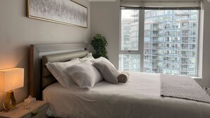 Una cama bien tendida con sábanas y almohadas blancas, una lámpara de noche y un cuadro abstracto enmarcado en la pared.