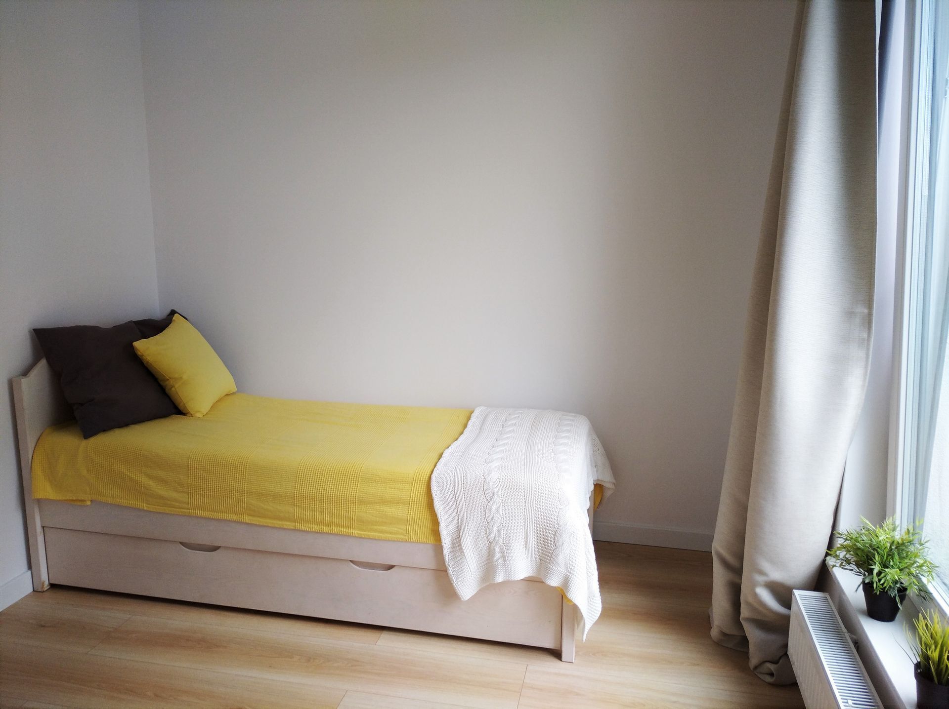 A single bed with a yellow blanket and a white throw, a grey pillow, and a white knitted throw. There is a window with curtains, a radiator, and a small potted plant on the windowsill.