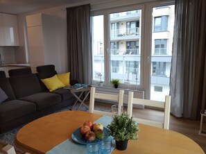 Ett modernt vardagsrum med en soffa, ett soffbord med äpplen och glas, ett litet bord med en växt och ljus, och en utsikt över en byggnad genom stora fönster.