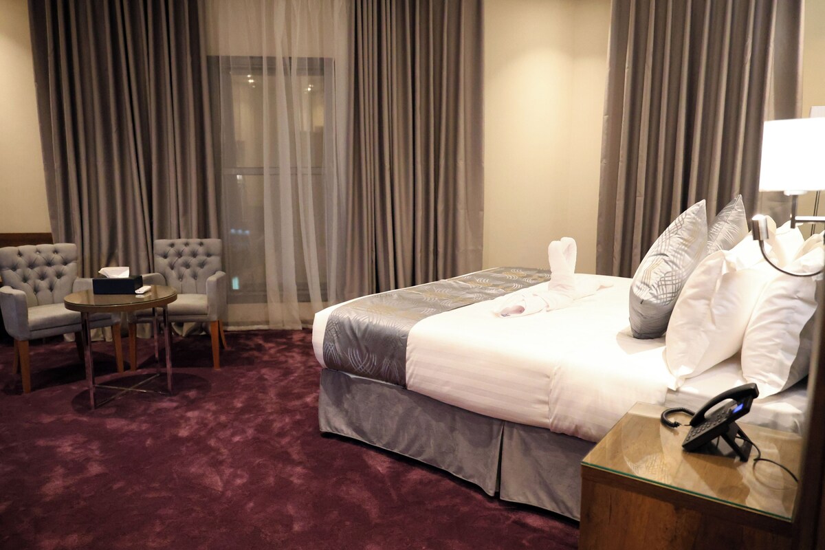 Ein Hotelzimmer mit einem Bett, einem Nachttisch mit Telefon, einem Sessel, einem kleinen Tisch mit Taschentuchbox und einem Fenster mit Vorhängen.