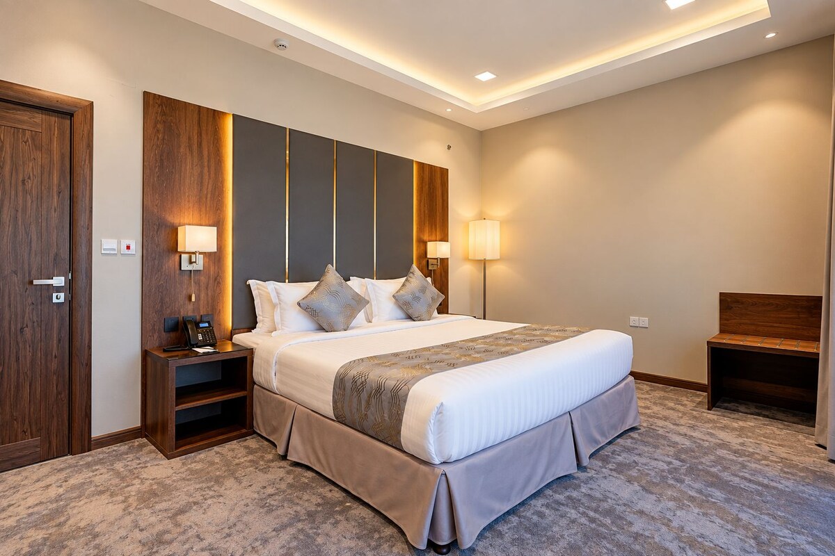 A hotel room with a large bed, bedside lamps, a desk, and a bench.