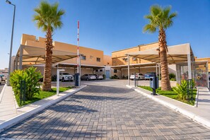 A parking area with multiple cars, palm trees, and a building with a flat roof. - Golden Season Hotel - AR Rass (Ar Rass)