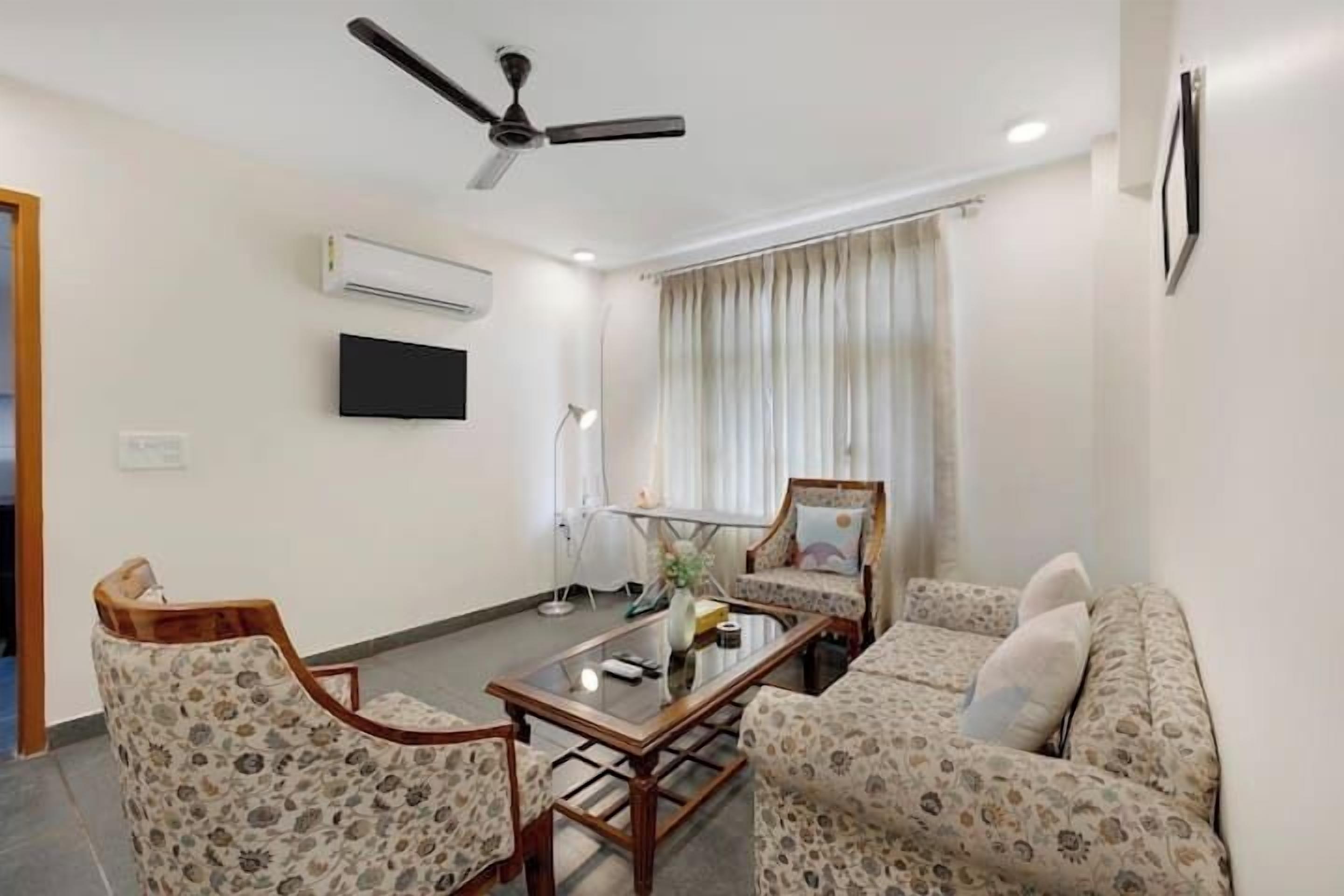 Una sala de estar con ventilador de techo, un televisor de pantalla plana, una mesa de centro de vidrio, sofás con estampado floral y un ventanal con cortinas.