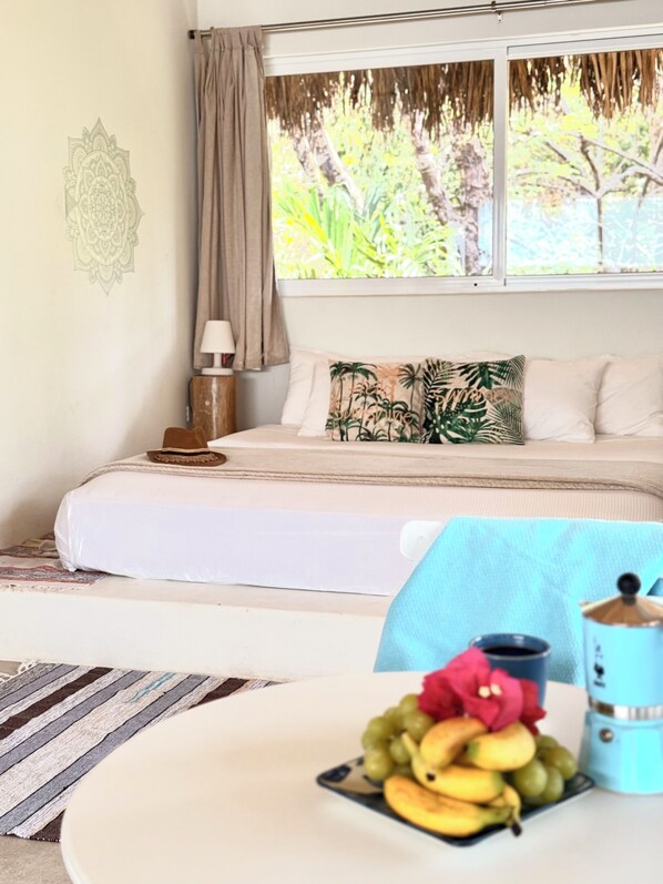 A bedroom with a bed, a nightstand, a lamp, a window with a view of trees, and a table with fruit and a container. - A natural setting in Playa Grande, peaceful and relaxing place! (Playa Grande)