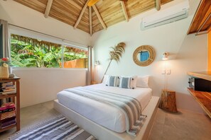 A bedroom with a large bed, a wooden ceiling, a window with a view of greenery, a round mirror, and a bedside table with books.